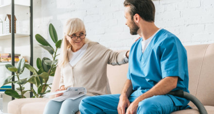 smiling senior woman sitting on couch and looking at social worker with vacuum cleaner