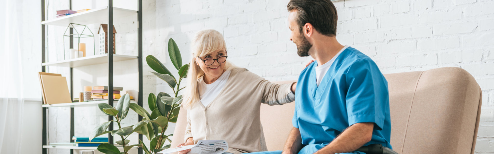 smiling senior woman sitting on couch and looking at social worker with vacuum cleaner